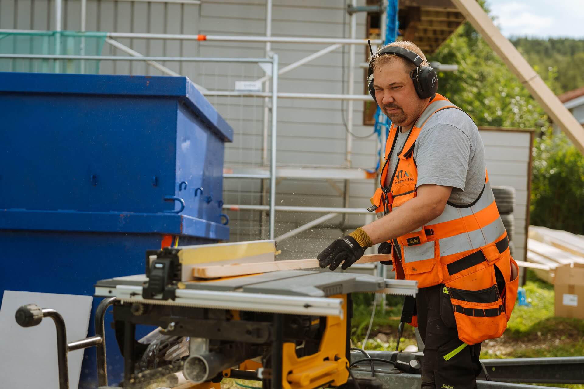 En mann med hørselvern som sager en planke med en sagmaskin utendørs.